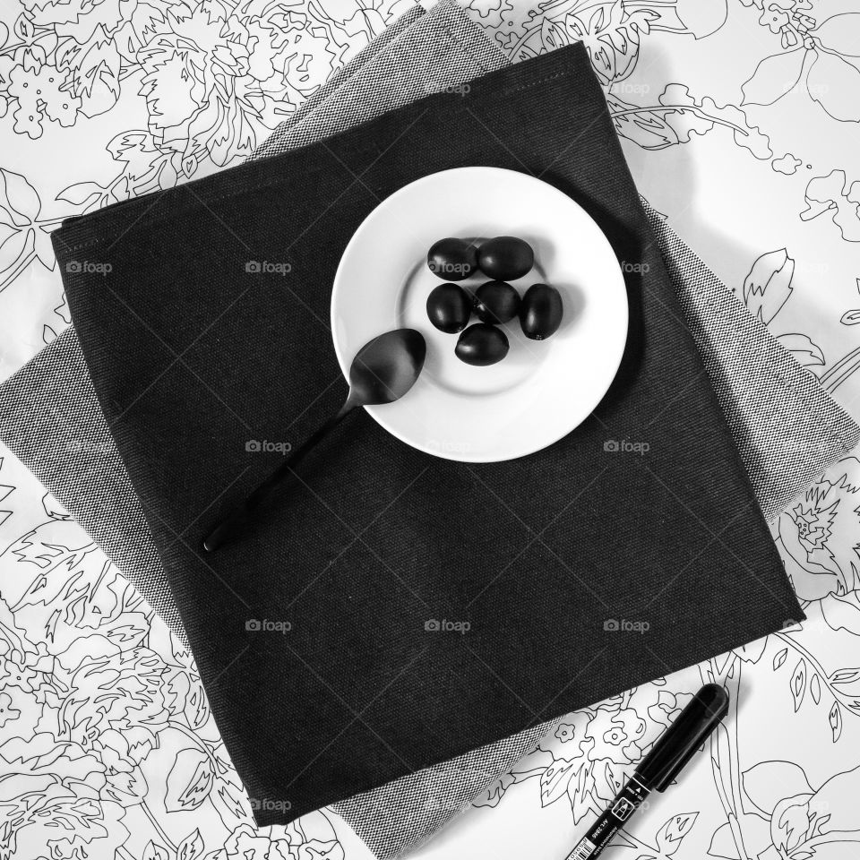 White saucer with black olives on a black and gray napkin on tracing paper with a floral graphic black and white pattern and marker. Workplace of the artist. Food photo, square orientation