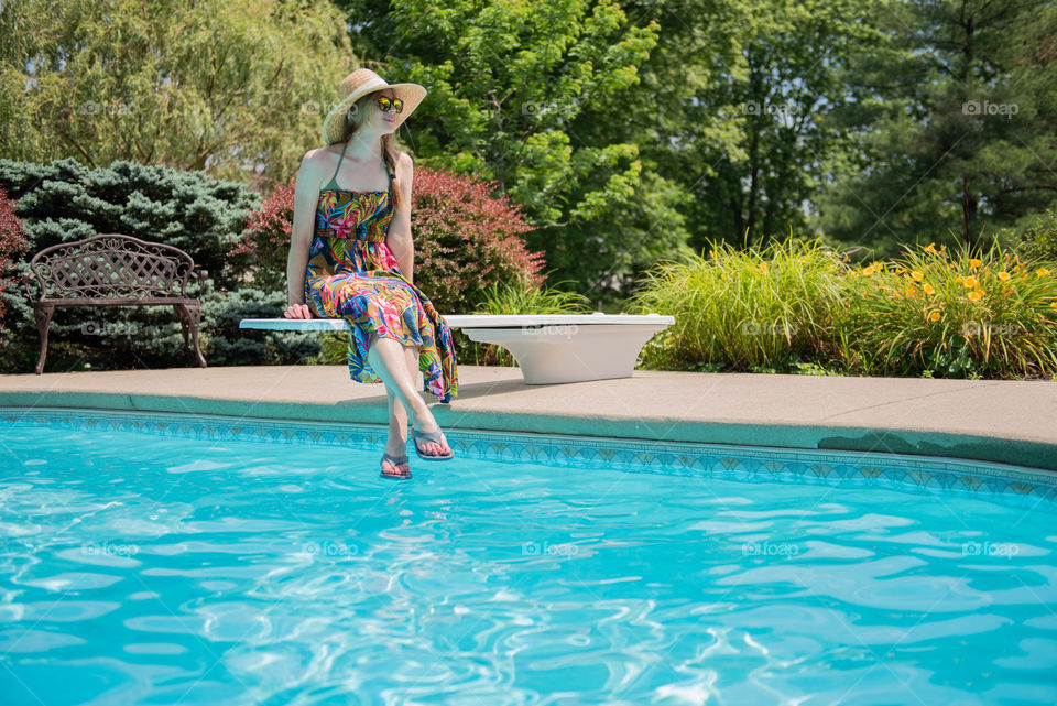 Young millennial woman wearing a dress and flip flops and sitting on the diving board of a swimming pool in the summer