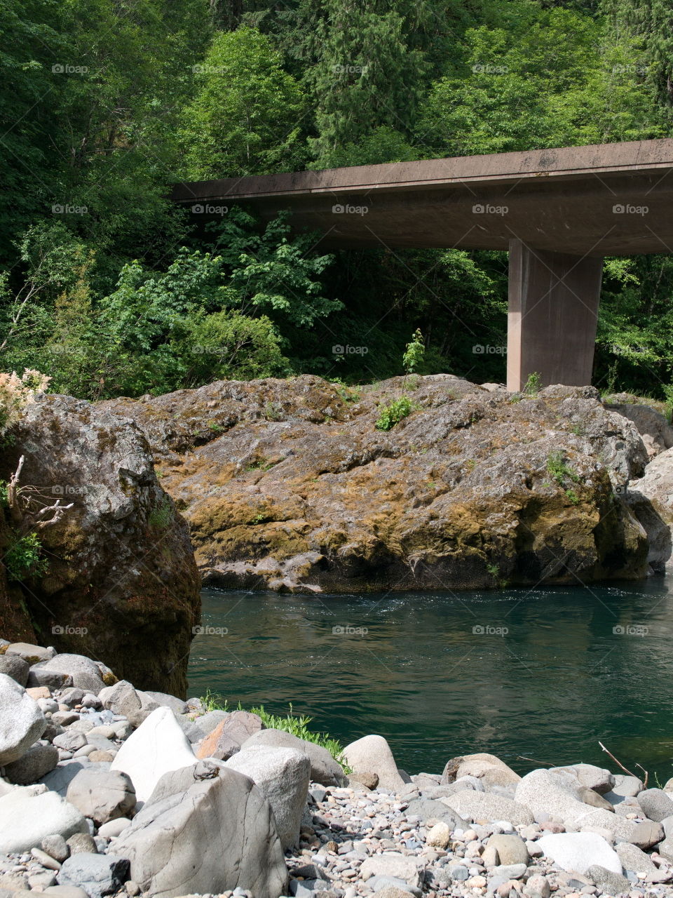 A small one-lane bridge crosses the magnificent waters of the Umpqua River and its boulder banks in Southwestern Oregon on a sunny summer morning. 