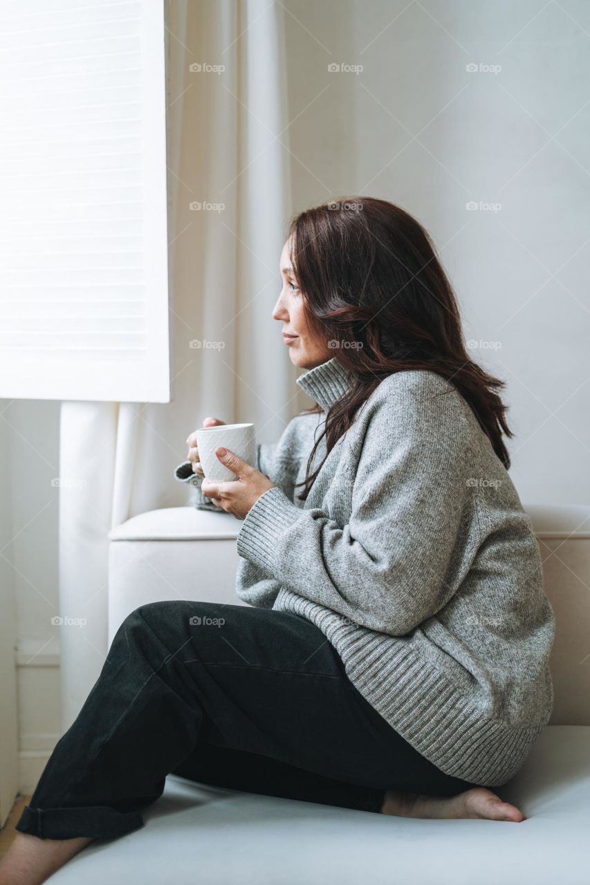 Thinking woman forty year with brunette long hair in cozy knitted sweater with cup of tea in hands in bright interior at home