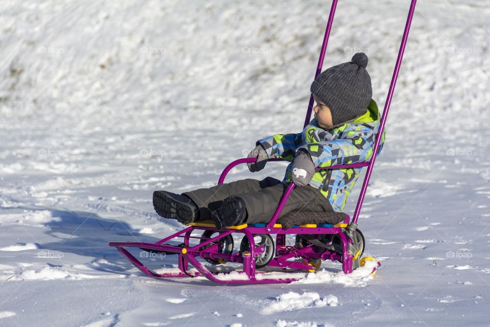 A child in winter in winter jackets, pants, a hat and boots on white snow on the street and in the park in nature plays winter fun and sleds.