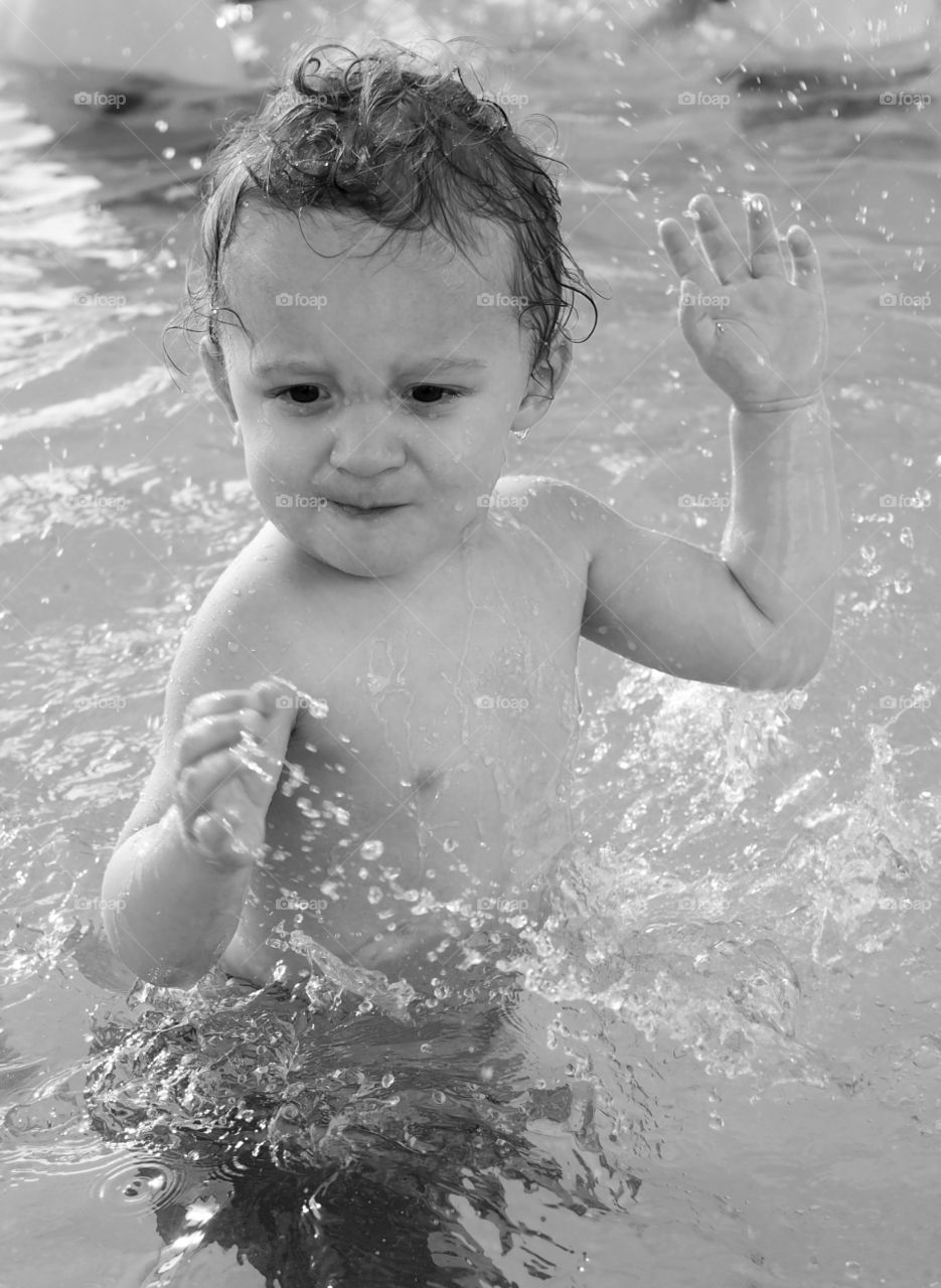 A small toddler boy splashing water in the shallow end of a swimming pool. 