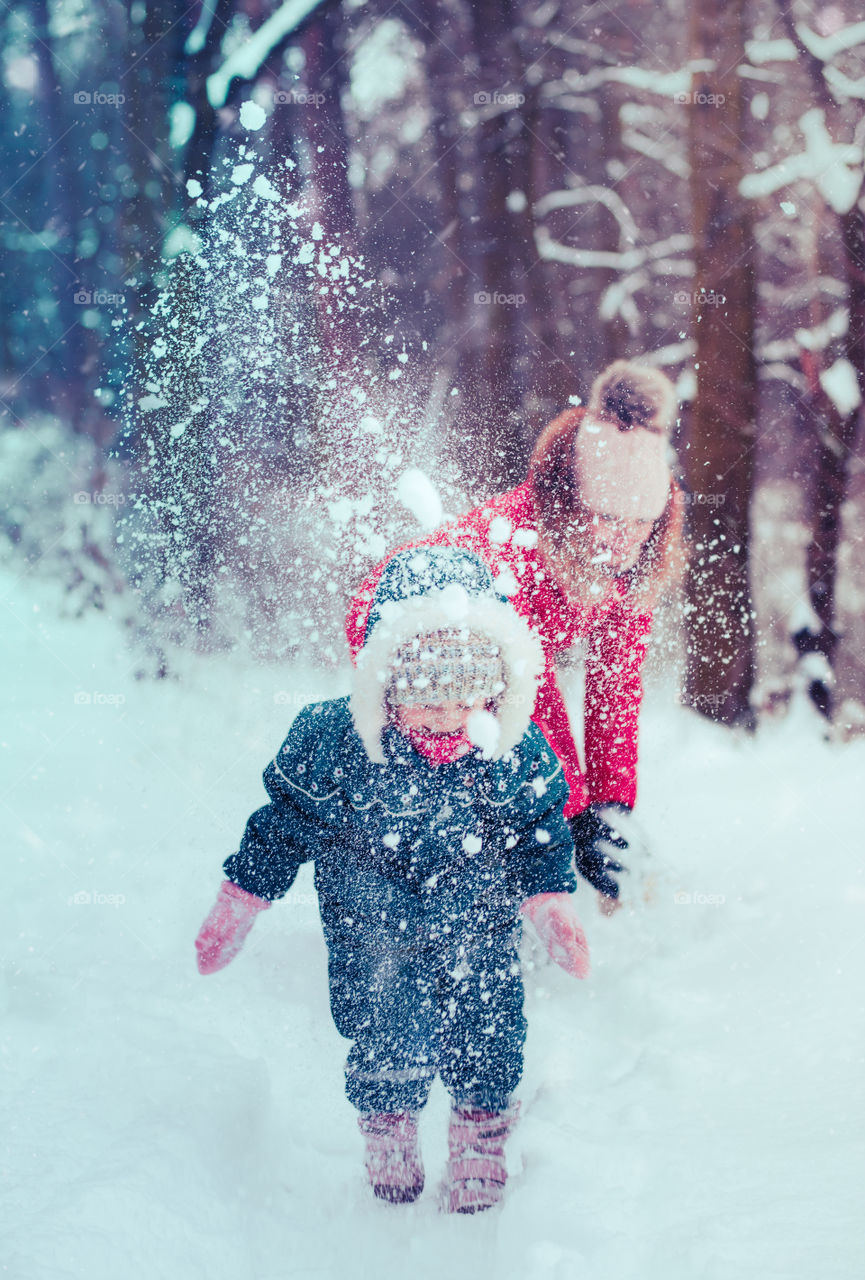 Mother is playing with her little daughter outdoors on wintery day. Woman is throwing snow on her child. Family spending time together enjoying wintertime. Woman is wearing red coat and wool cap, toddler is wearing dark blue snowsuit