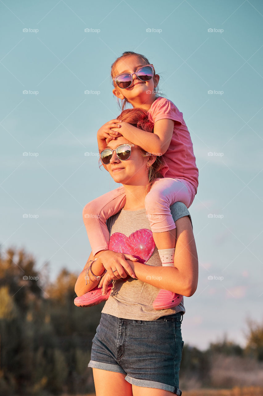 Sisters enjoying piggyback ride. Teenage girl carrying her younger sister up on the back and shoulders spending time playing together outdoors in the countryside. Candid people, real moments, authentic situations