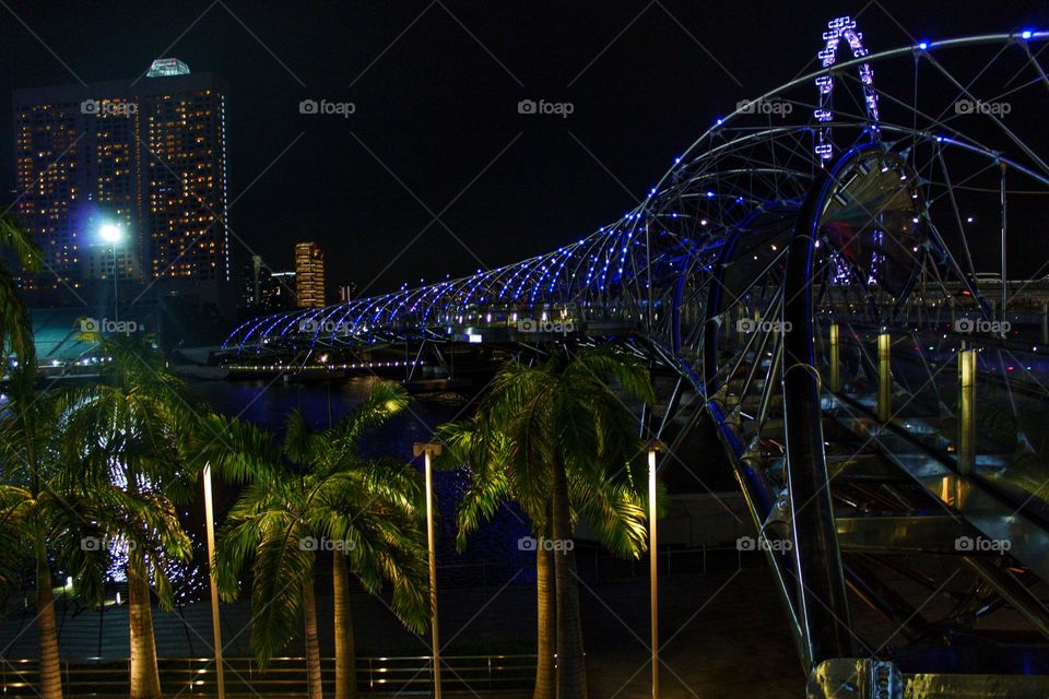 Illuminated helix bridge with palm trees, ferris wheel and tall building at night in Singapore.