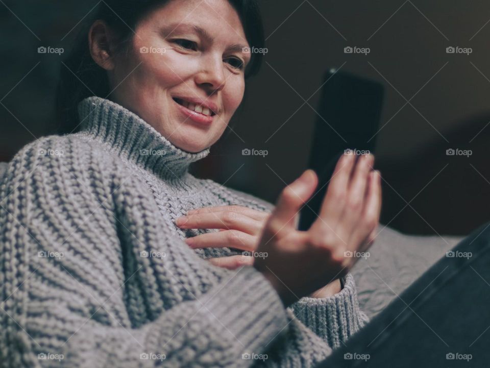 Portrait of one beautiful Caucasian brunette girl in a gray sweater sitting on the floor by the bed and holding a smartphone in her hands and communicating via video conference with a happy emotion on her face, bottom side view close-up.