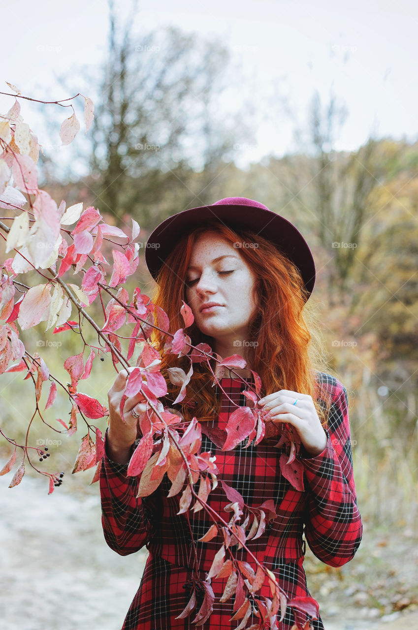 Beautiful portrait of young redhead curly woman standing near tree with red leaves in colorful autumn park in red hat and dress. Fall season.