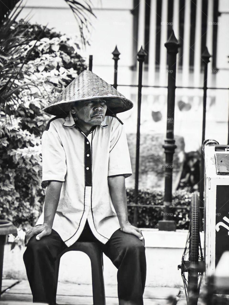 Black and white photo portrait showing the atmosphere of the city. People sitting on benches wear straw hats and carts selling food. The background of the photo is an iron fence and plants which provide color contrast