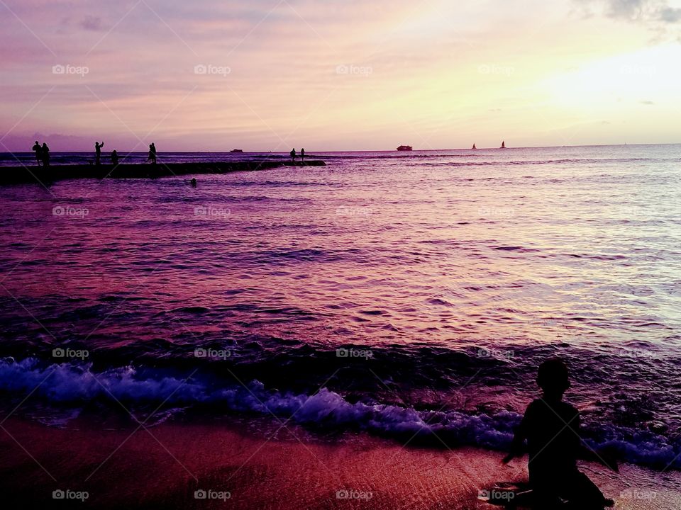 boy plays on Waikiki Beach as the sun sets and pants the ocean beautiful colors of purple pink and gold