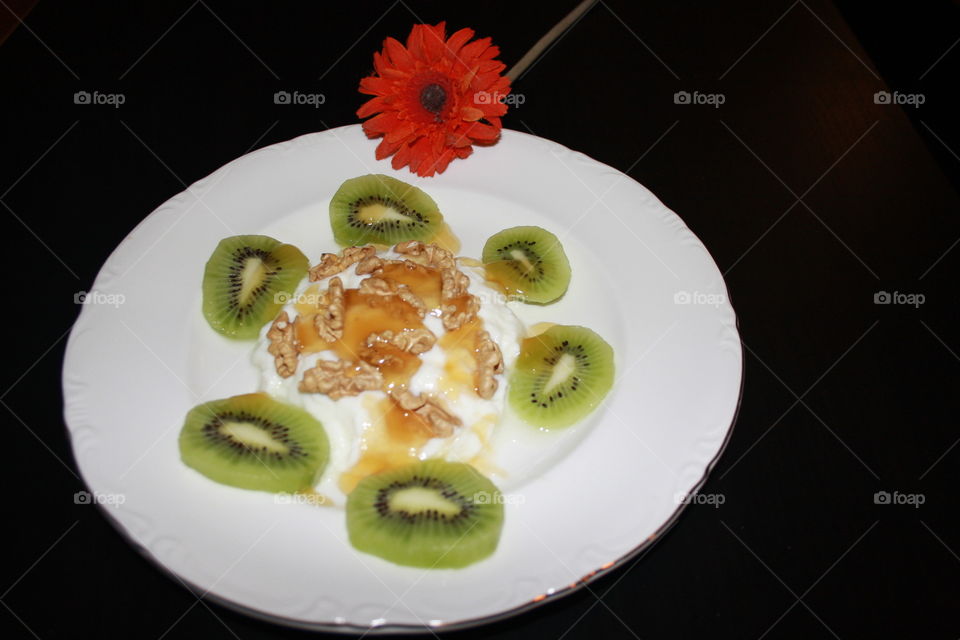 There is yogurt on the plate and on top of the yogurt I have added walnuts and honey. Around the yogurt are kiwi slices. On the plate is a flower.