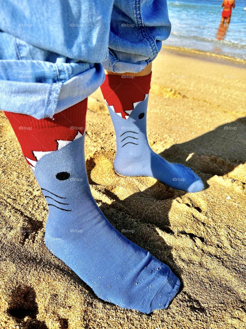 A selfie foot shot of me in my shark attack socks standing in the damp sand at the edge of the Pacific Ocean with a woman in a red long distance swim suit entering the water in the background 