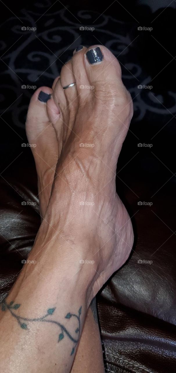Feet of a woman or girl with ankles crossed. She is relaxing with legs elevated sitting in her brown leather recliner. Black toenail polish is drying.