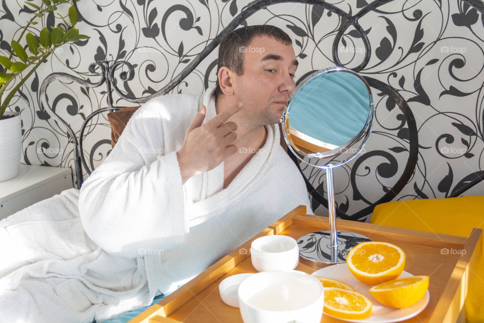 A man and home spa care at home takes care of himself and smears his face with cream, does a manicure at home on a blue background and with oranges and rests.