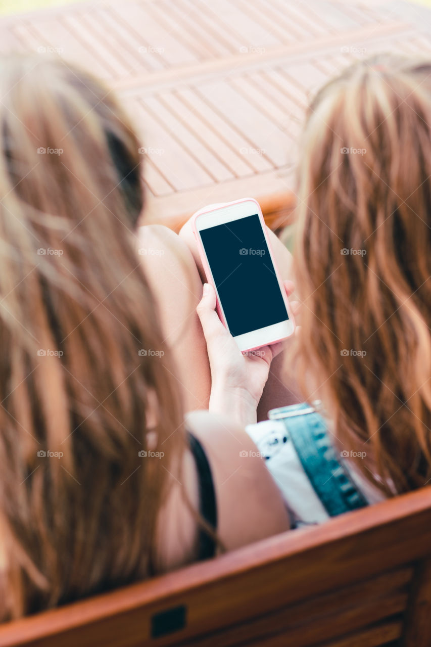 Young women using mobile phones watching music clip, texting, messaging. Teenagers using the smartphones, sitting on bench outdoors. Using technology devices. Girls wearing summer clothing