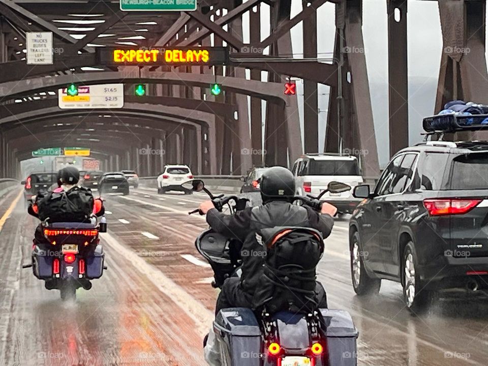 Motorcyclists riding over a bridge in traffic on wet roads in rainy weather with an expect delays warning sign