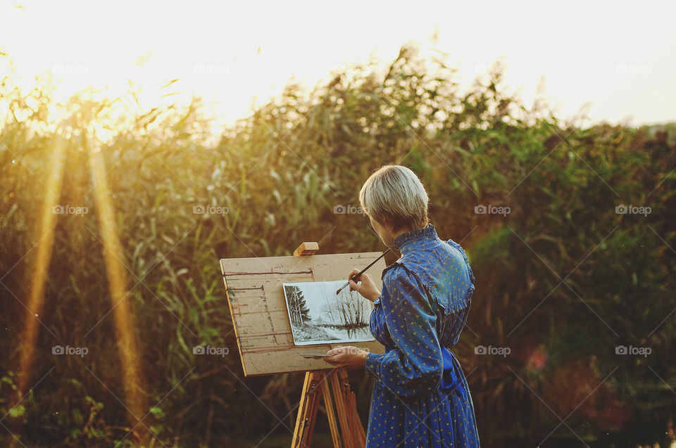 Lovely young woman artist in blue dress paints her picture in autumn at sunset light in nature.