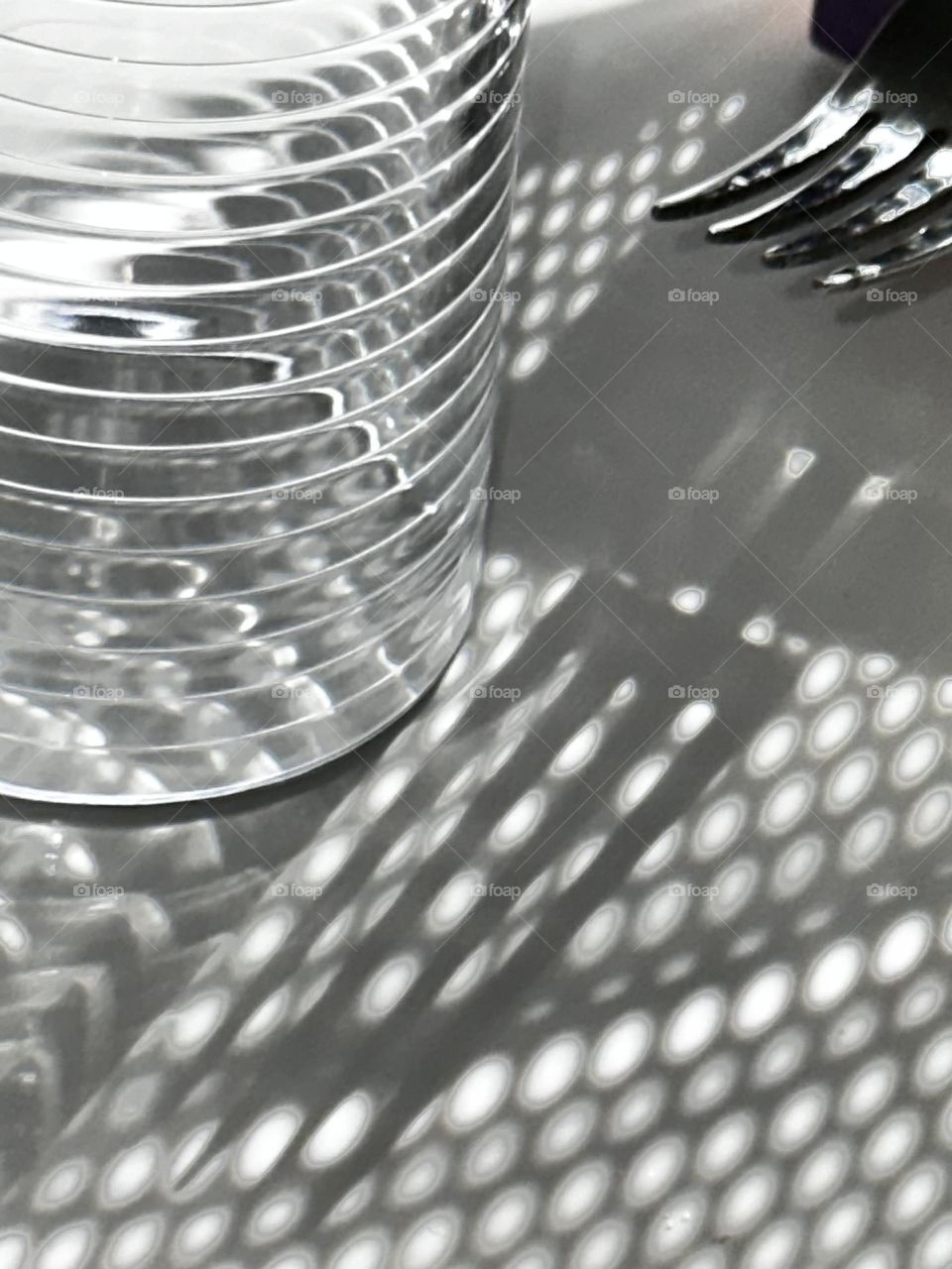 A stainless steel dinner fork casting a shadow under a metal colander next to a drinking glass with a ring pattern