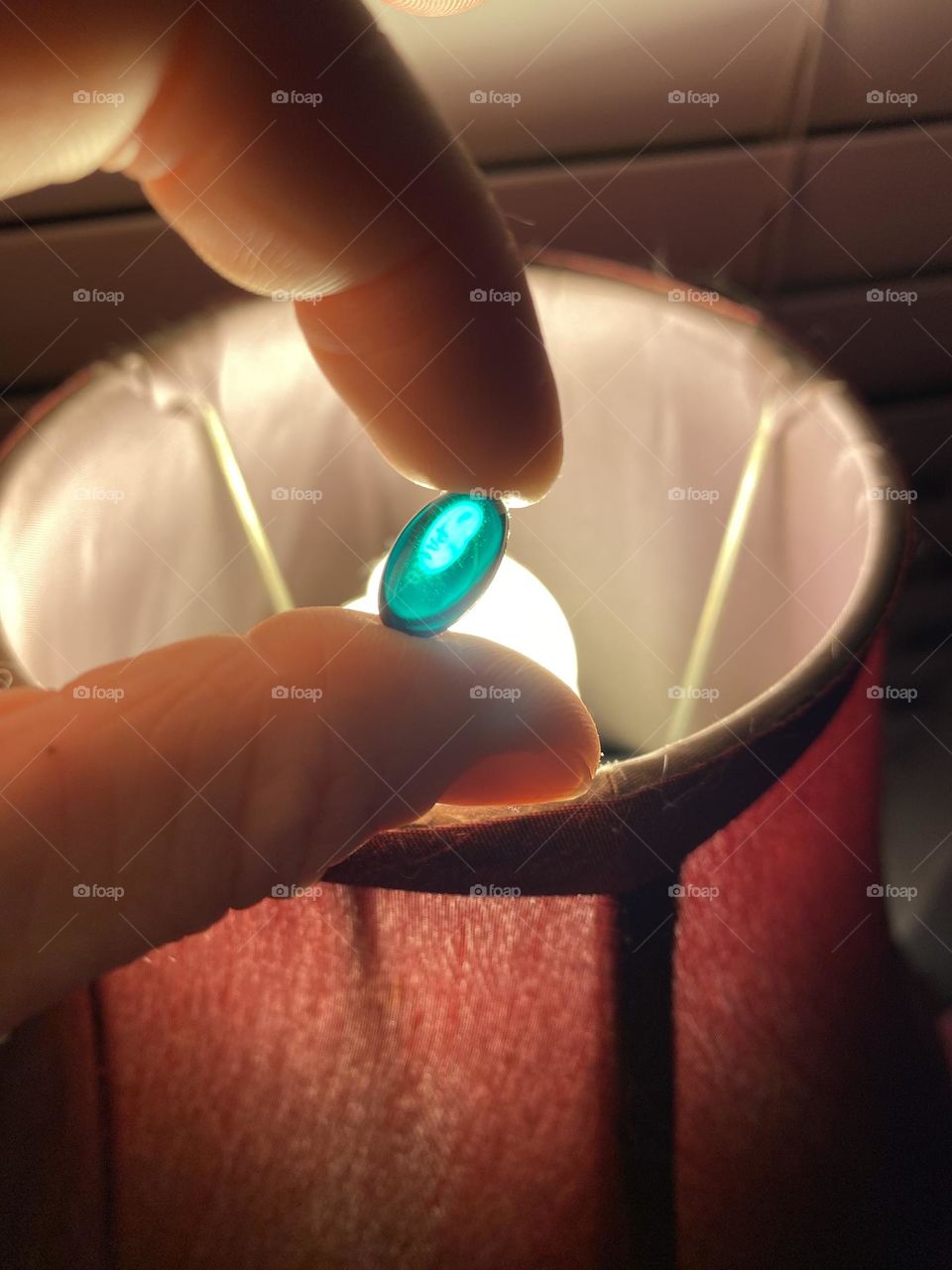 A blue-green pill is held between two fingers in the light of the lamp. 