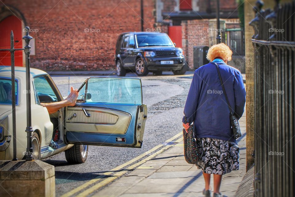 Nostalgia. An old lady walks past a vintage car with the owner propping her feet up on the door.