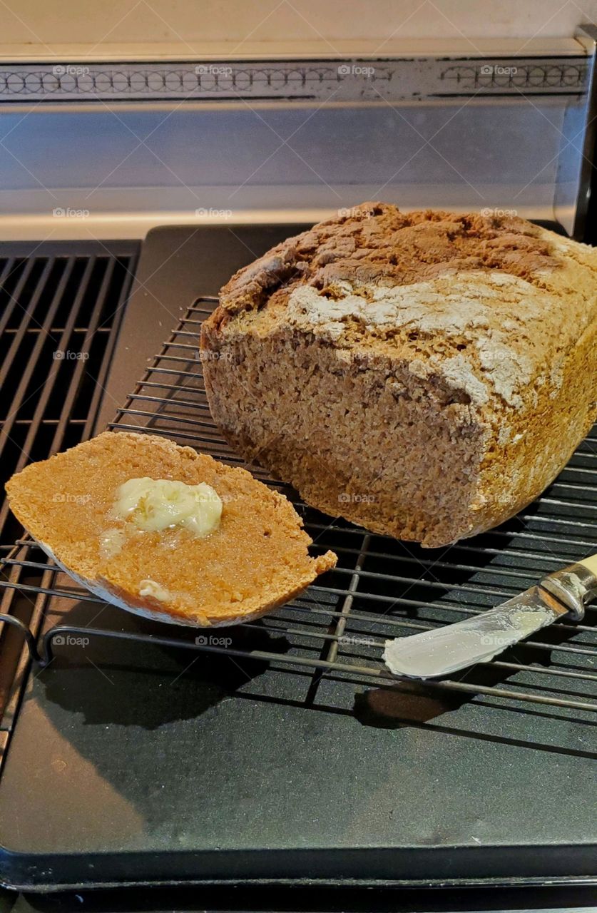 Fresh loaf of homemade bread cooling on rack along with end piece of bread with butter melting on it. There's a butter knife next to loaf as it cools.