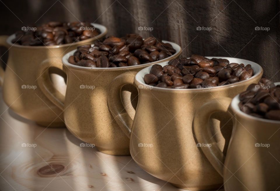 A row of 4 coffee cups, filled with coffee beans against a wood background with shadow lines across them 