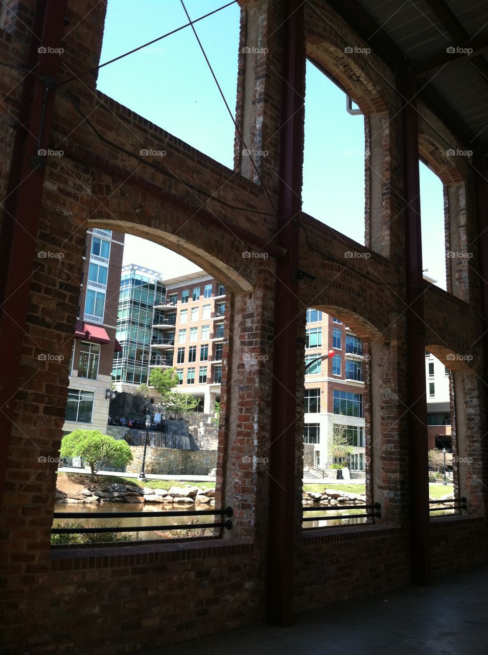 Modern buildings along river walk seen through arched windows of historic brick building.