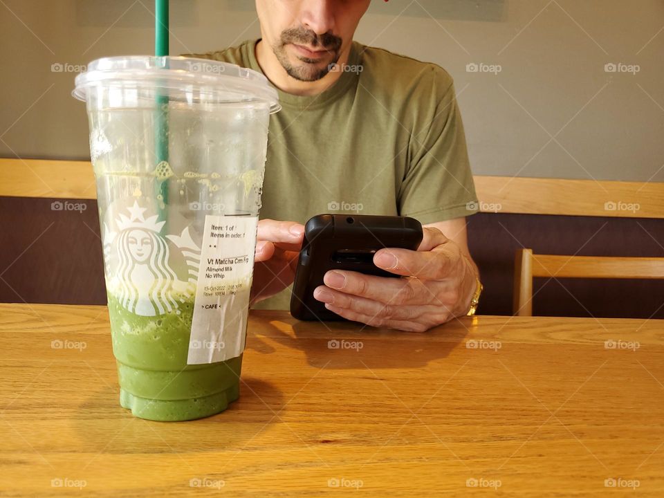 A man on his cell phone while sitting inside Starbucks with a matcha frappuccino.