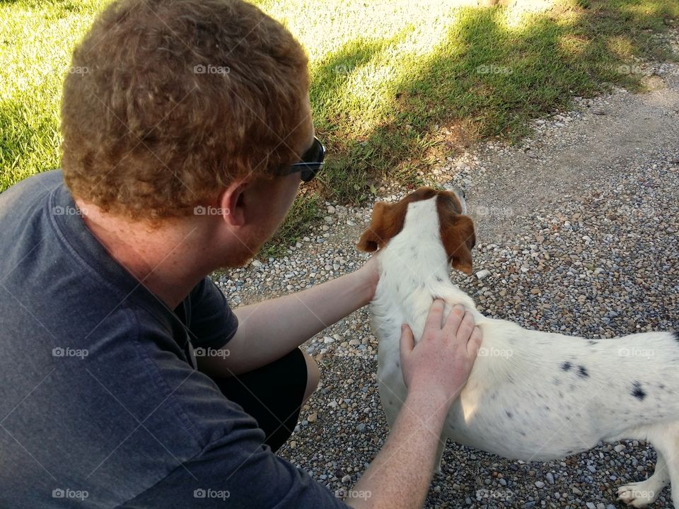 A red headed man petting a dog on a gravel road with green grass in spring