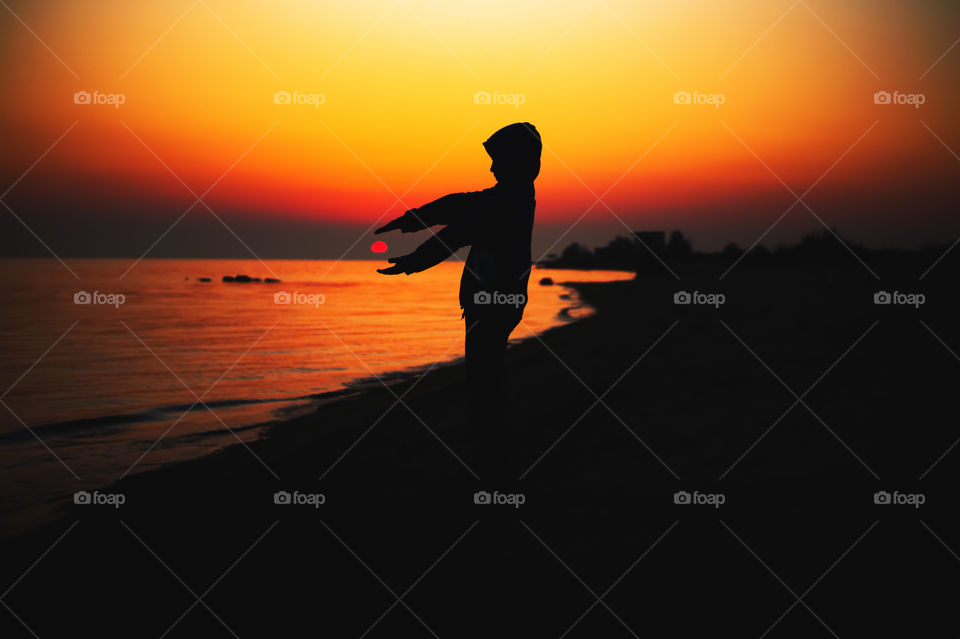 Silhouette of a boy holding the sun in his hands at sunset, against the background of the sea