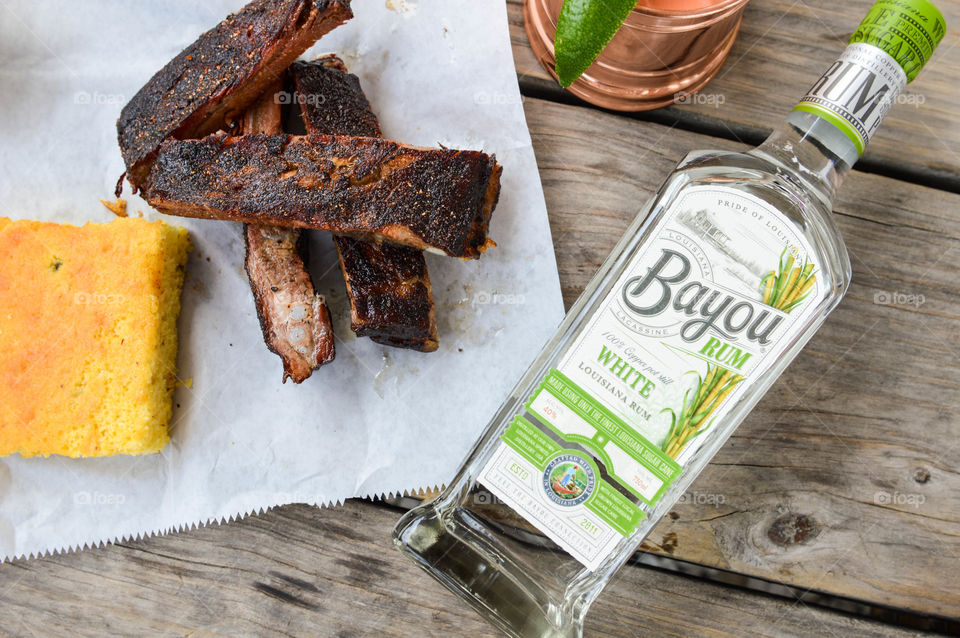 Bottle of Bayou white rum laid out on a wooden table with a copper mug and smoked ribs