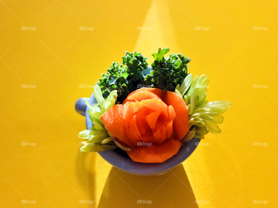My amateur attempt at an orange carrot rose nestled together with dark green curly kale & pale green flower petals in a tiny blue teacup on a bright yellow background. The early evening sunlight produced a triangular light beam & shadow. 
