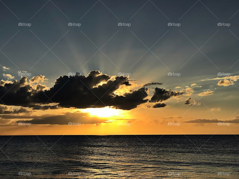 Golden sunset behind intense dark clouds emitting bright sun rays over the ocean.