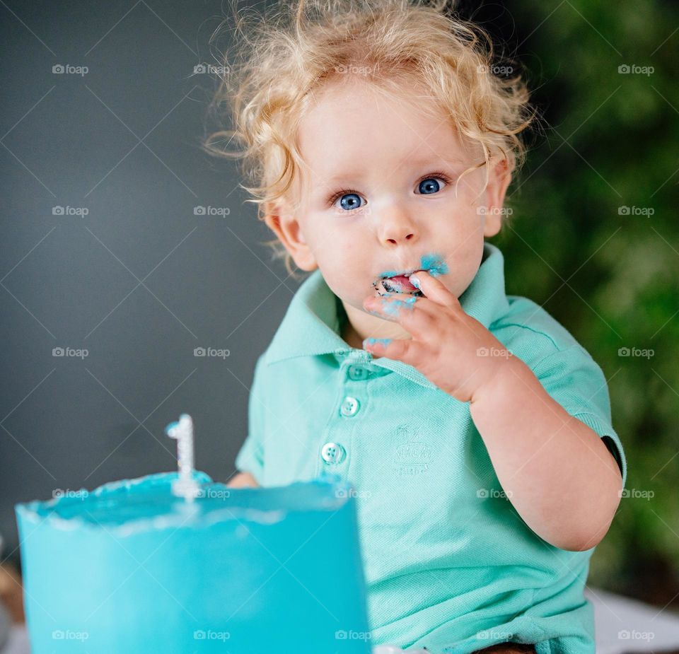 Cute little boy in blue t-shirt eating cake. Blond curly hair and blue eyes