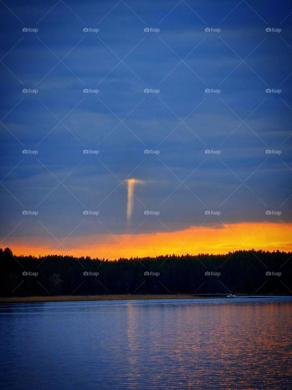 Sunset on the lake. Trees on the shore of the lake. The whole sky is covered with dark clouds. Only above the trees id a yellow sunset. A ray of light breaks through dense clouds