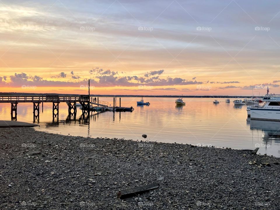 A peaceful summer sunset begins over Hampton Harbor in Seabrook, NH, casting a golden glow along the horizon as calm waters reflect the soft colors of the evening sky.