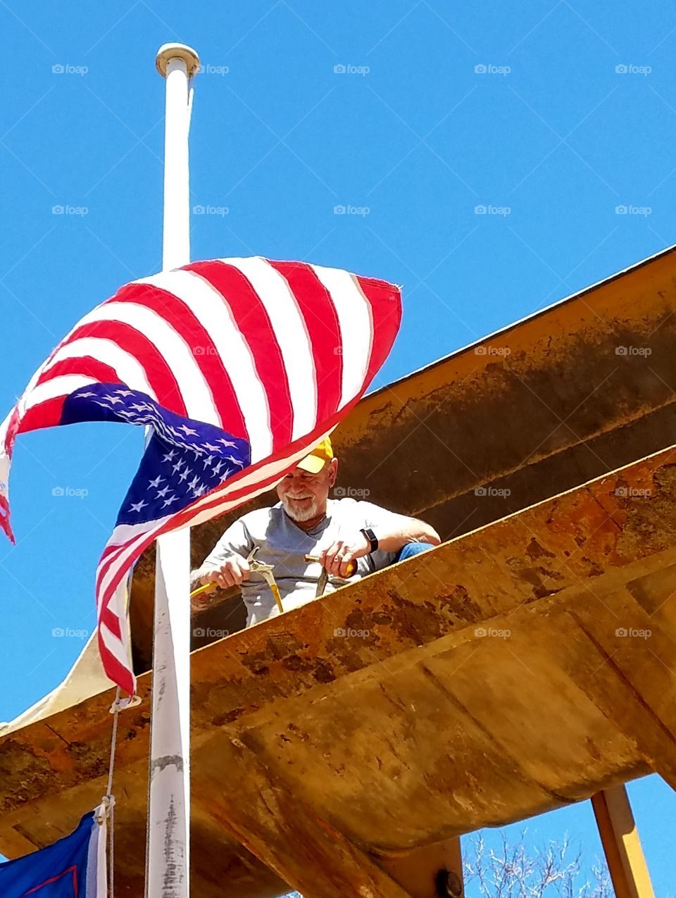 American flag🇺🇸blowing in wind as  Worker in Bucket of Front End Loader assembles solar light for top of flagpole. He's 20 feet above ground to reach pole top. Tools in hands work to put light together.