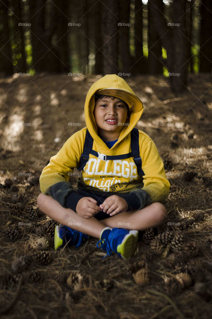 Seasonal outdoor portrait of a young happy Eurasian kid hiking in a pine forest wood.Natural setting, the boy is wearing blue sneakers, yellow sweater, hood and cap, smiling while looking at the camera, seating on the floor surrounded by pinecones.
