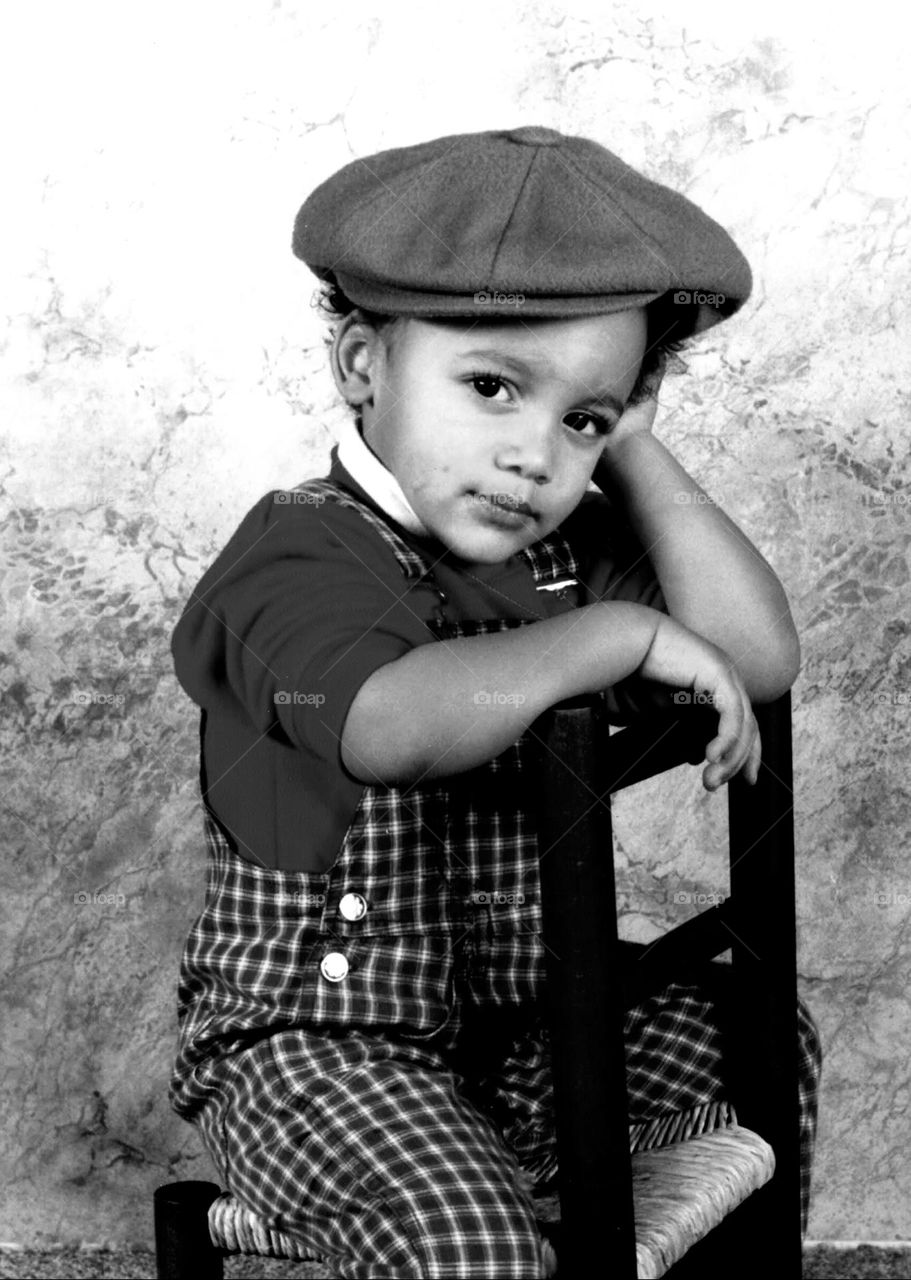 Vintage black and white shot of a little boy wearing a newsboy cap sat astride on a straw chair