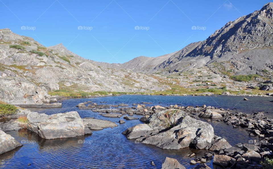 A blue sky and colorful tundra set the stage for an enormous mountain and high alpine lake to shine and delight. Ripples in the water add interest and texture.