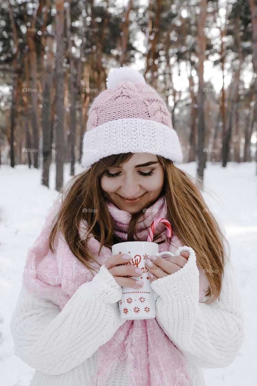 Girl in warm clothes holding cup and looking to cup in winter forest