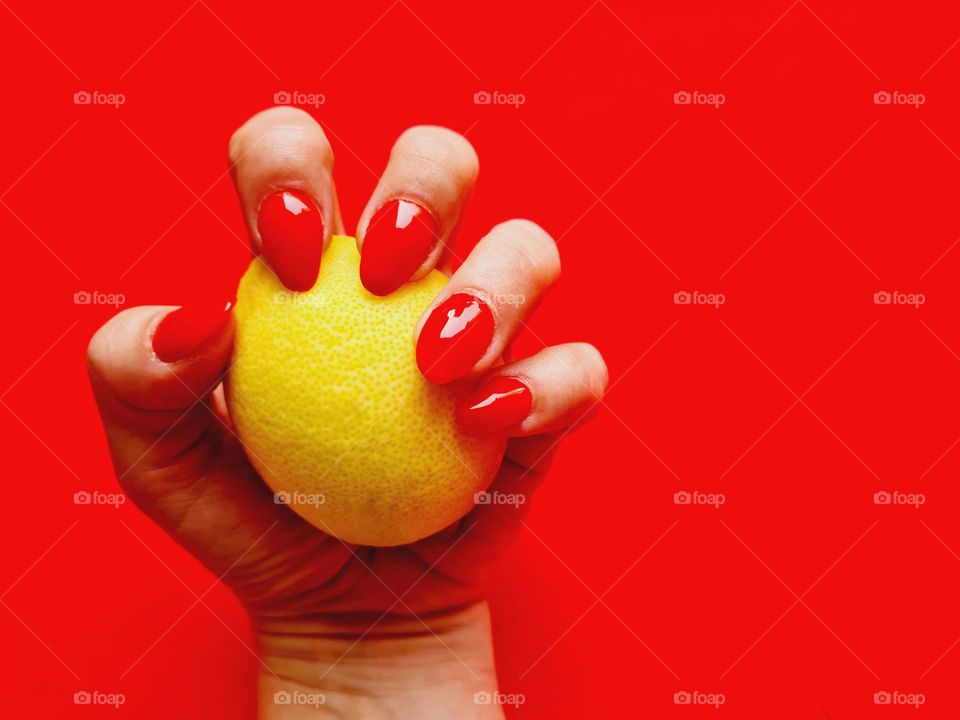 hand with red lacquered nails squeezes a yellow lemon on a red background