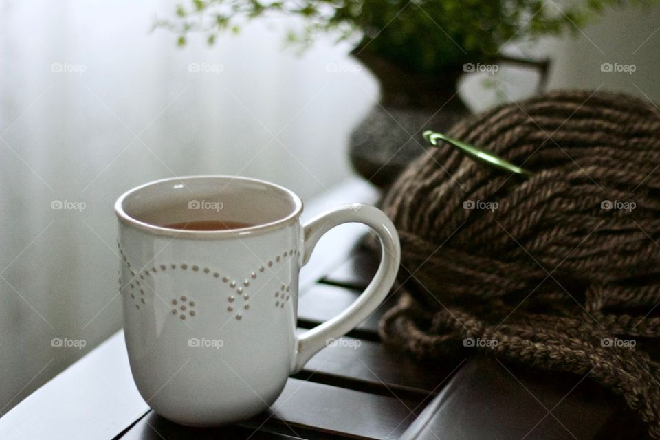 Hot tea in a white mug, yarn and crochet hook on a wooden surface, small plant in a pitcher and white curtain in the blurred background