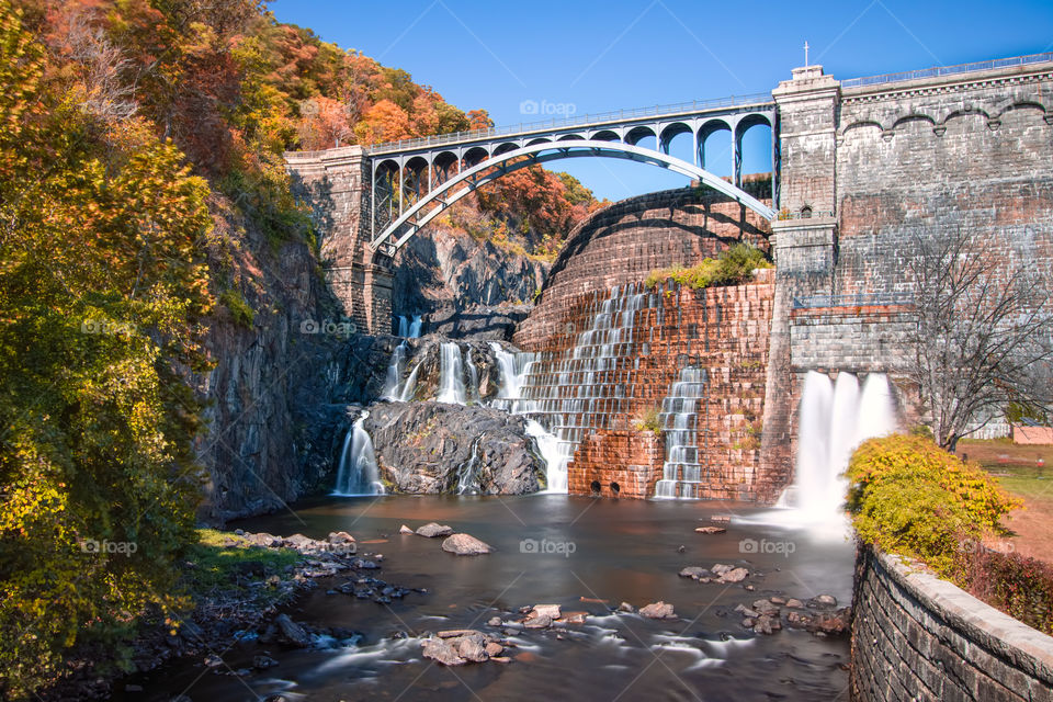 Beautiful waterfall under a clear blue sky, surrounded by colorful leaves as fall foliage begins to appear in autumn. 