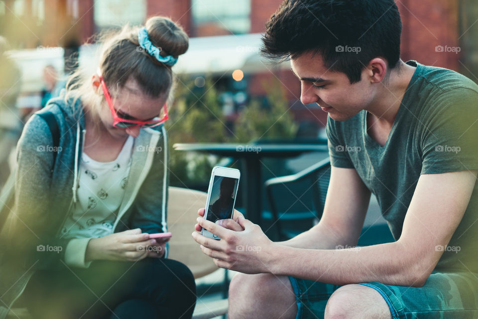 Couple of friends, teenage girl and boy, having fun using smartphones sitting in center of town, spending time together