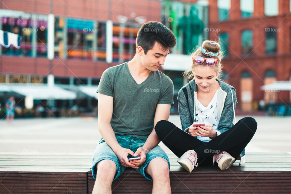 Couple of friends, teenage girl and boy, having fun together, using smartphones, sitting in center of town, spending time together. Real people, authentic situations