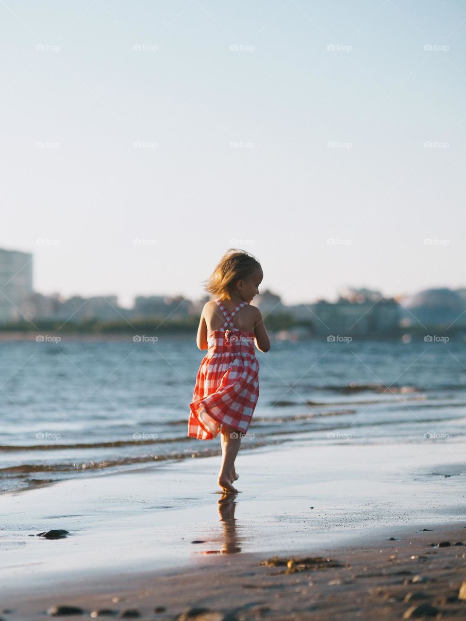 Cute little girl with short blonde hair wearing red dress walking on seaside in sunny summer day