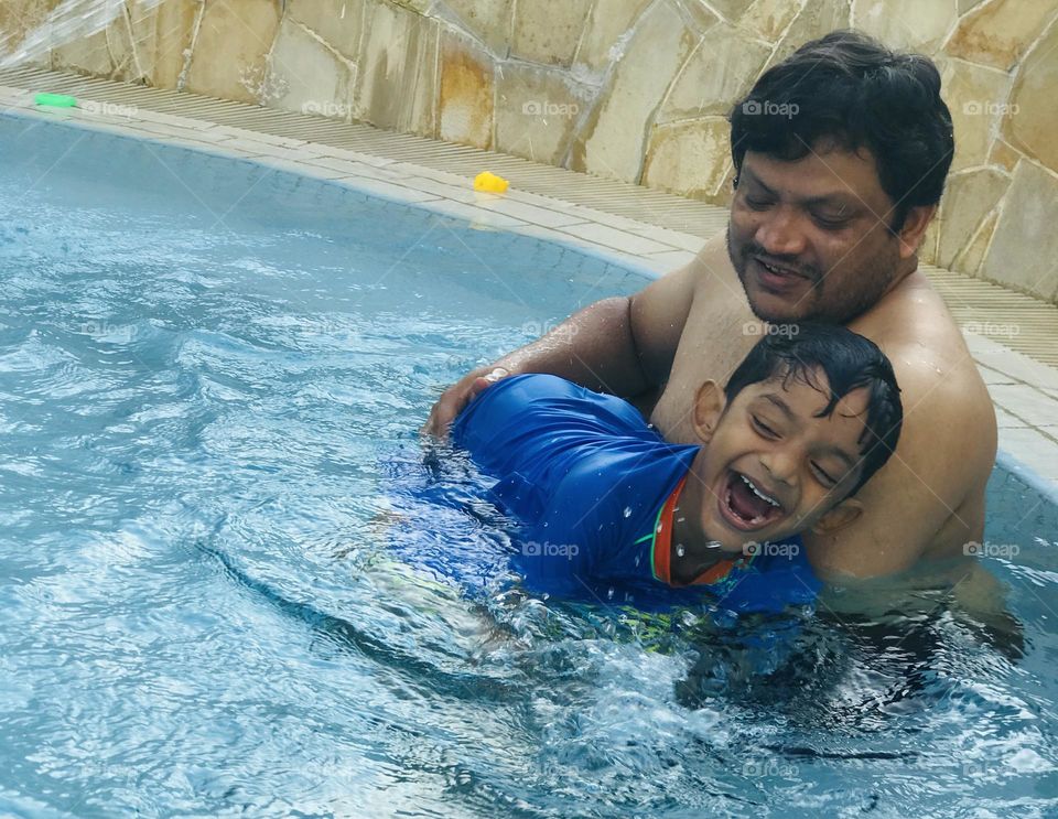 A father and his son both spending time at swimming pool at summer and father holds son on his hands ,and the boy is enjoying and smiling like anything.