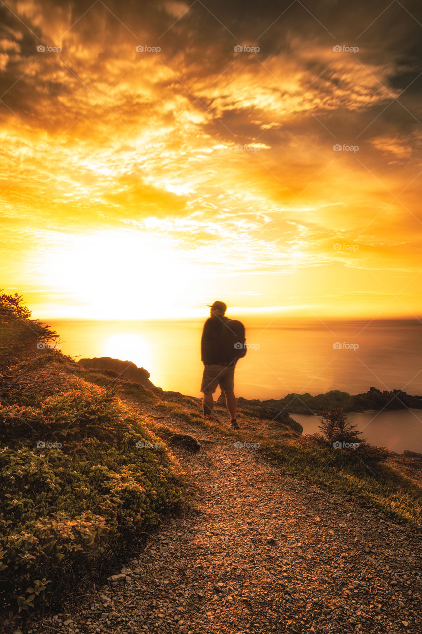 Silhouette of a male hiker walking along a cliff overlooking the ocean, as the setting sun casts beautiful golden light onto the landscape. 