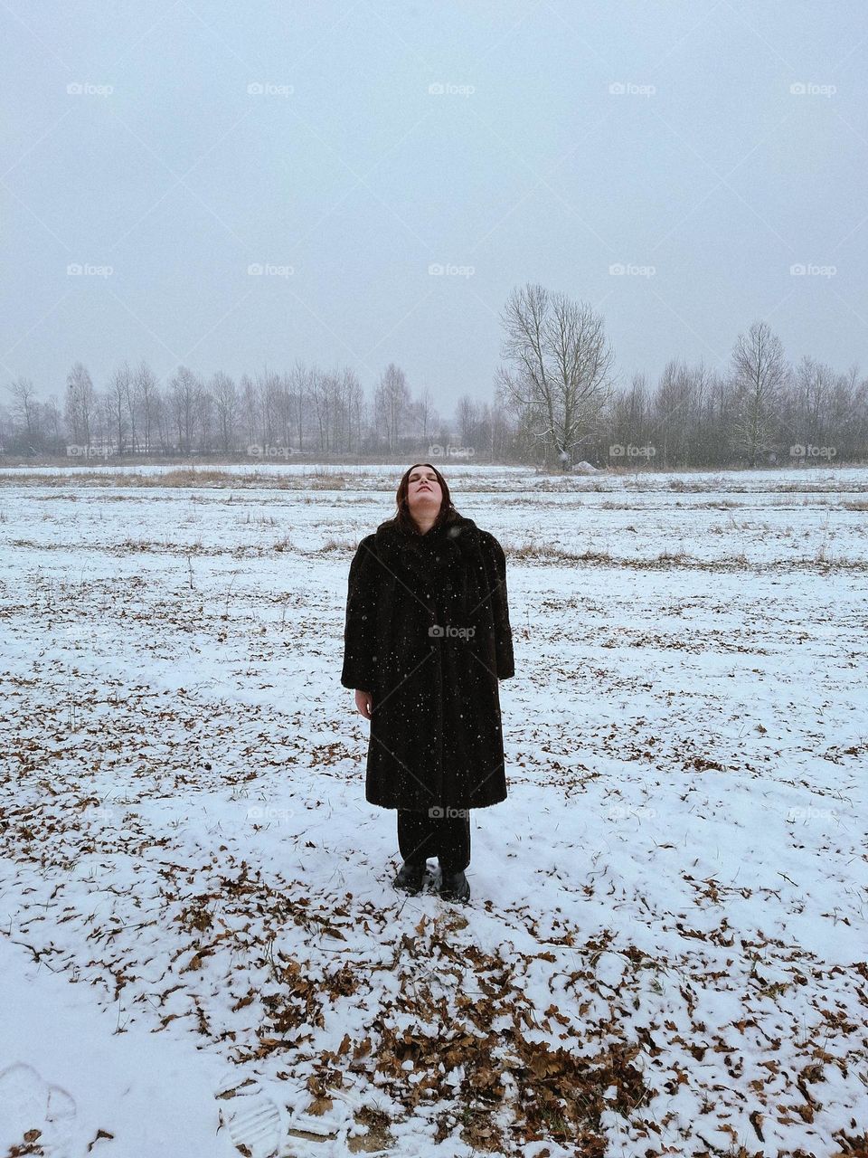 Winter photo of a girl standing in the middle of a wild snowy field, enjoying the snowflakes