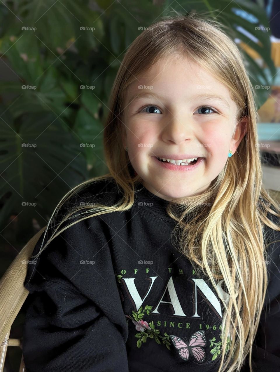 happy joyful blonde girl with pierced ears, rocking her turquoise earrings and VANS sweatshirt in front of a monstera plant